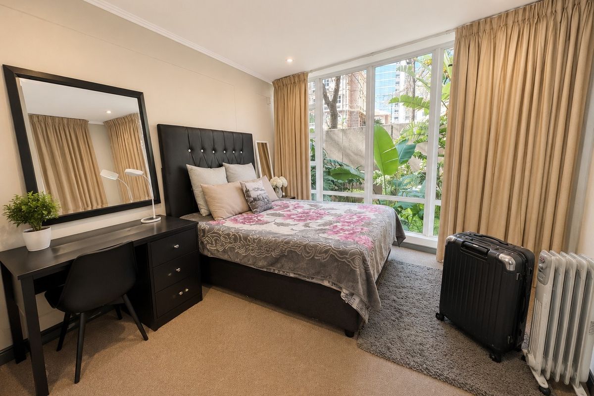 A well-lit bedroom featuring large floor-to-ceiling windows with garden views, neutral carpeting, and recessed ceiling lighting.