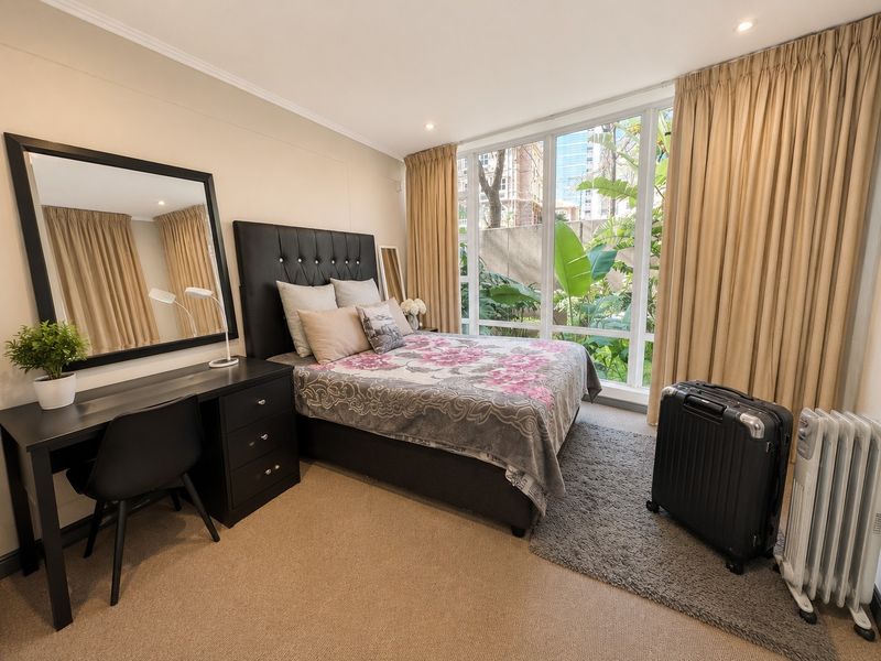 A well-lit bedroom featuring large floor-to-ceiling windows with garden views, neutral carpeting, and recessed ceiling lighting.