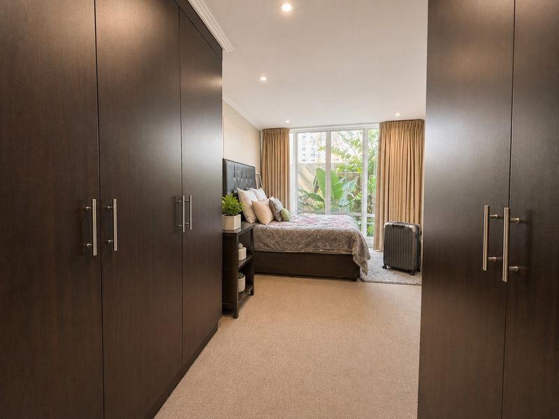 A well-lit bedroom featuring extensive built-in cupboards, neutral carpeting, and a large window providing natural light and a view of greenery.