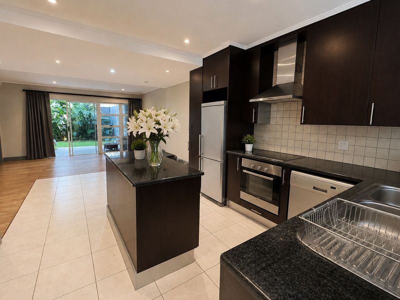 Modern open-plan kitchen featuring dark wood cabinetry, granite countertops, and integrated appliances, flowing seamlessly into a bright living area with garden access.