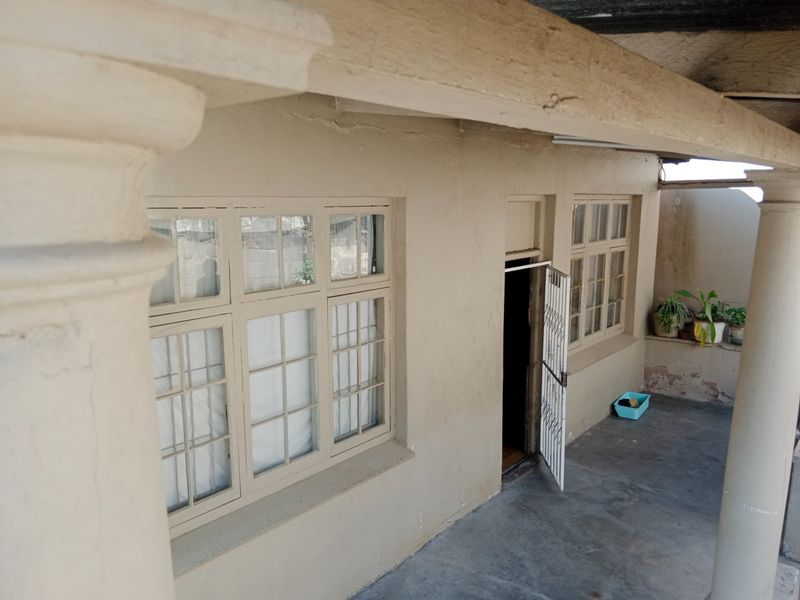A covered patio area featuring concrete columns, multi-pane windows, and a security gate entrance, showing signs of wear on the exterior wall and flooring.