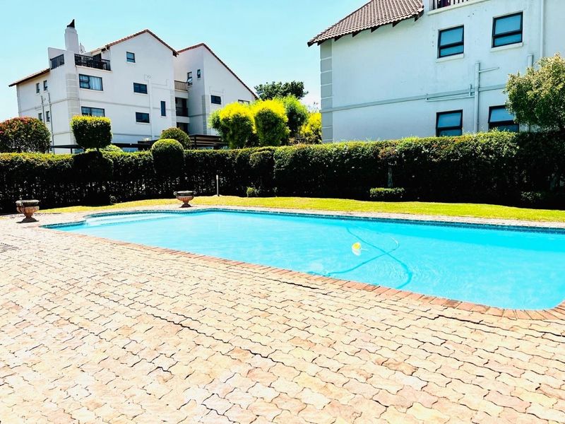 A well-maintained communal swimming pool area featuring a paved deck and landscaped hedges, set within a residential apartment complex.