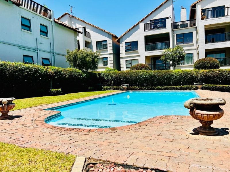 A well-maintained communal swimming pool area surrounded by paved decking, manicured hedges, and multi-story apartment buildings with balconies.