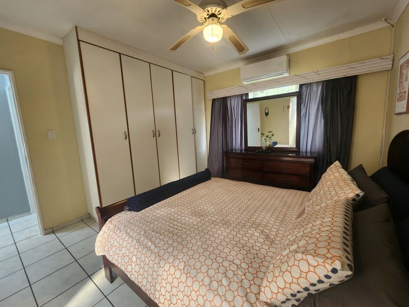 A functional bedroom featuring built-in cupboards, a ceiling fan, and a wall-mounted air conditioning unit with tiled flooring throughout.