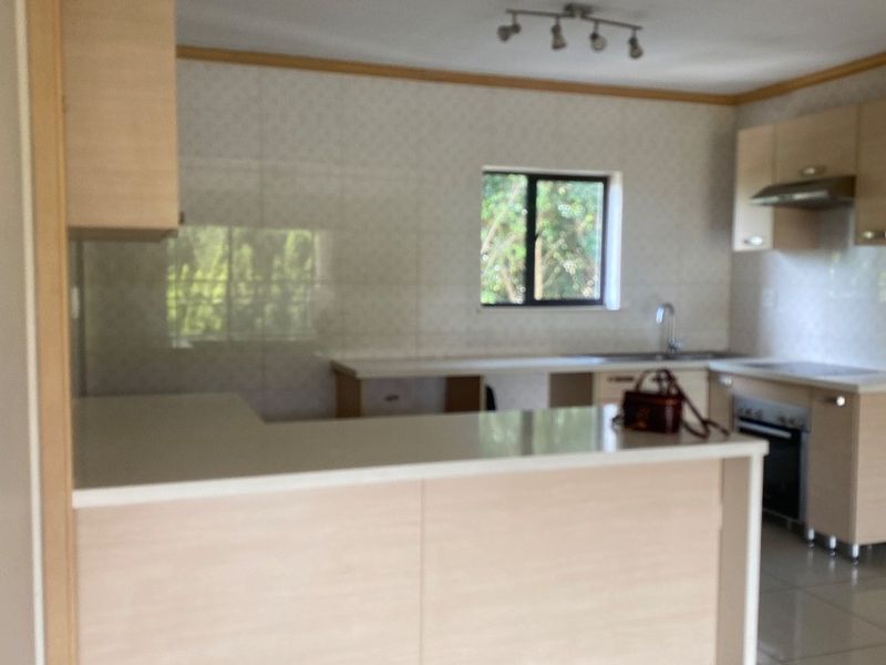 A clean, modern kitchen featuring light wood cabinetry, stone countertops, built-in appliances, and neutral tiled flooring in an open-plan layout.