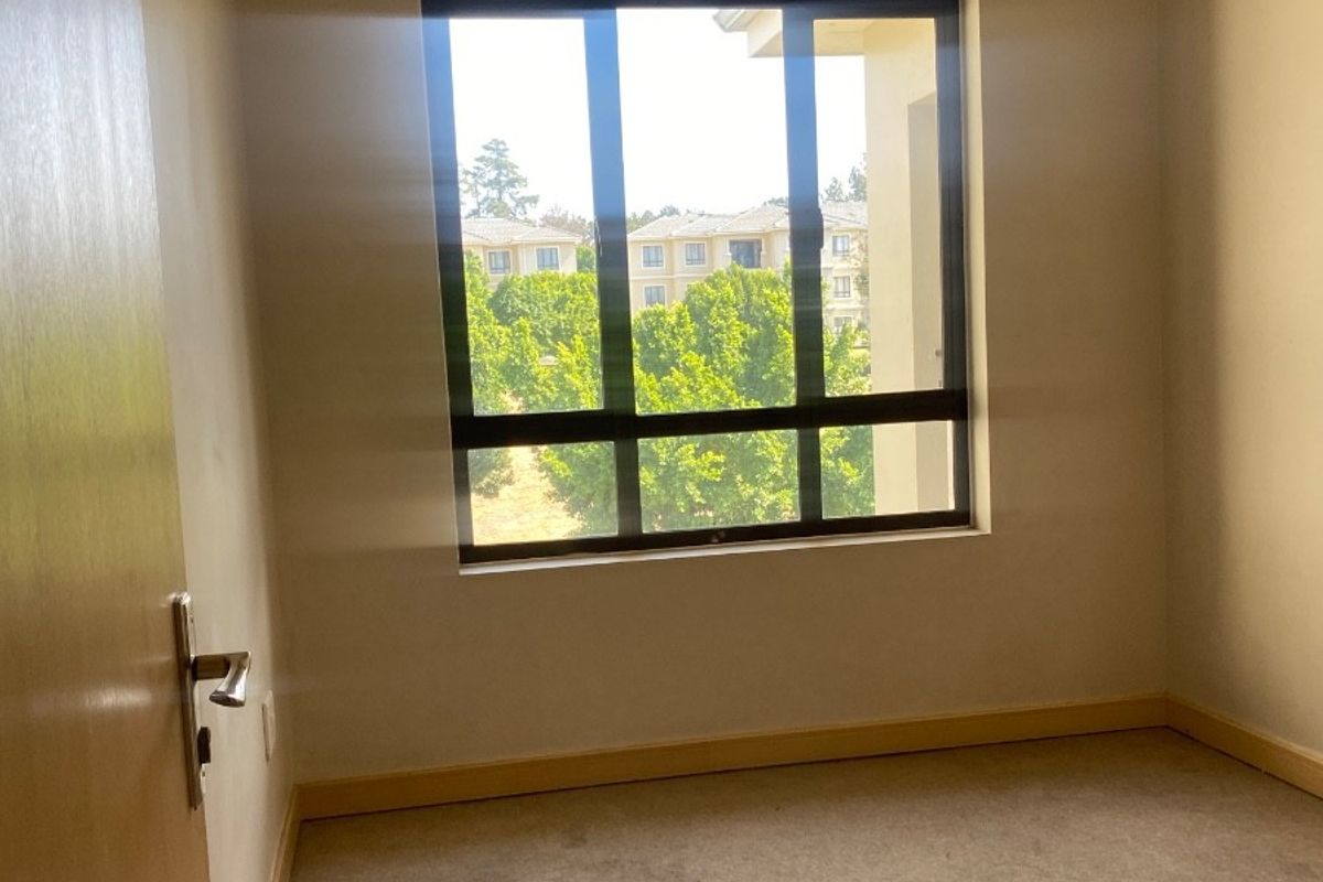 An empty bedroom featuring neutral carpet flooring, timber trim details, and a large window providing natural light and a view of the surrounding complex.