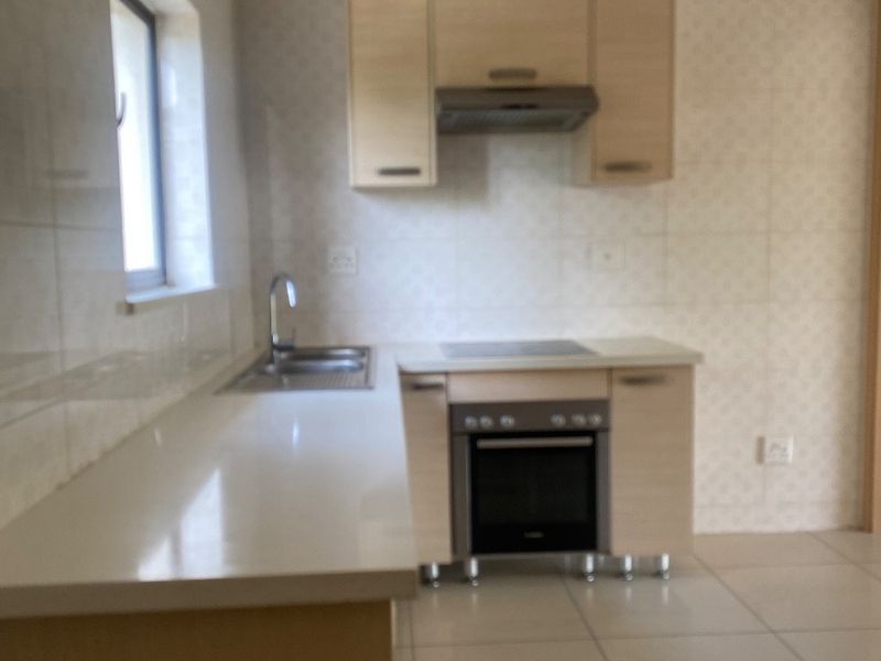 A compact, functional kitchen featuring light wood-finish cabinetry, built-in oven and hob, stainless steel sink, and neutral tiled flooring.