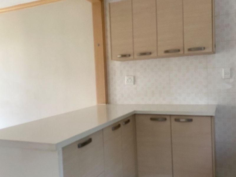 A clean, empty kitchen featuring light wood-tone cabinetry, white stone countertops, and neutral tiled flooring with a functional open-plan layout.