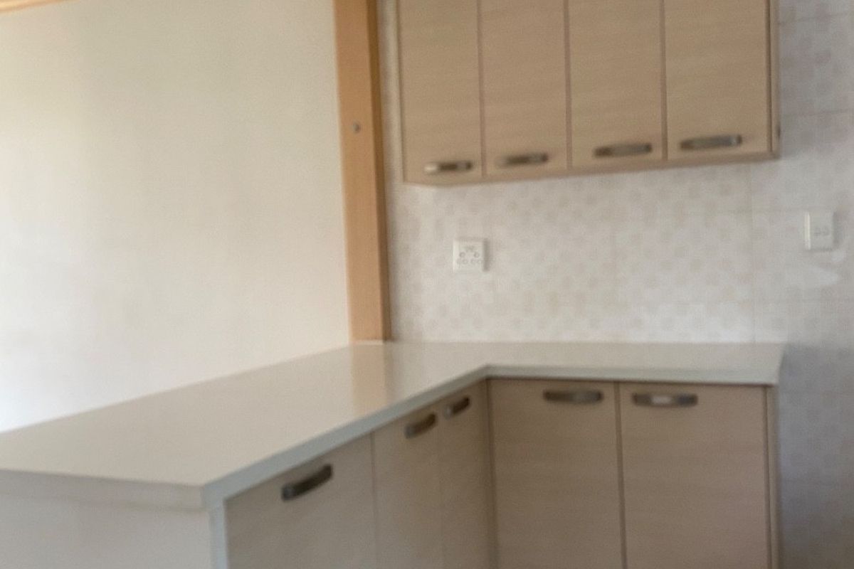 A clean, empty kitchen featuring light wood-tone cabinetry, white stone countertops, and neutral tiled flooring with a functional open-plan layout.