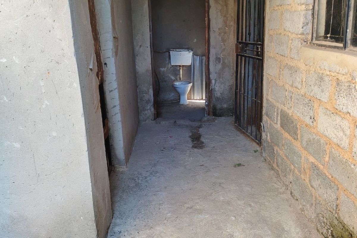 A basic outdoor bathroom facility featuring a toilet, concrete flooring, and exposed brick walls in need of significant renovation and repair.