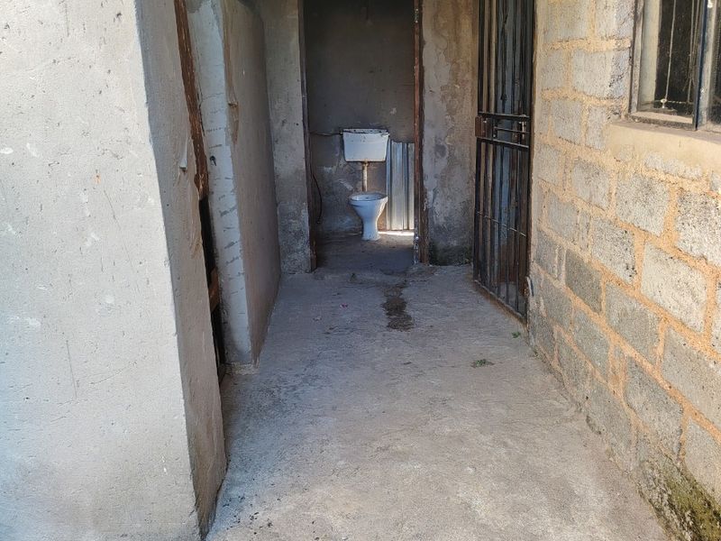 A basic outdoor bathroom facility featuring a toilet, concrete flooring, and exposed brick walls in need of significant renovation and repair.