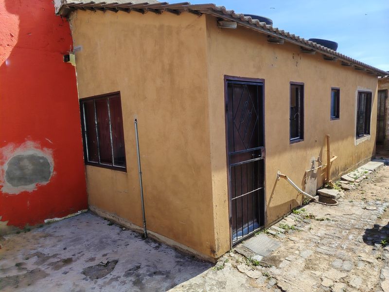 Rear exterior view of the property featuring a paved yard, security gates, and metal window frames, showing signs of wear and maintenance requirements.