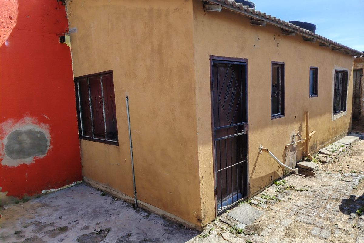 Rear exterior view of the property featuring a paved yard, security gates, and metal window frames, showing signs of wear and maintenance requirements.