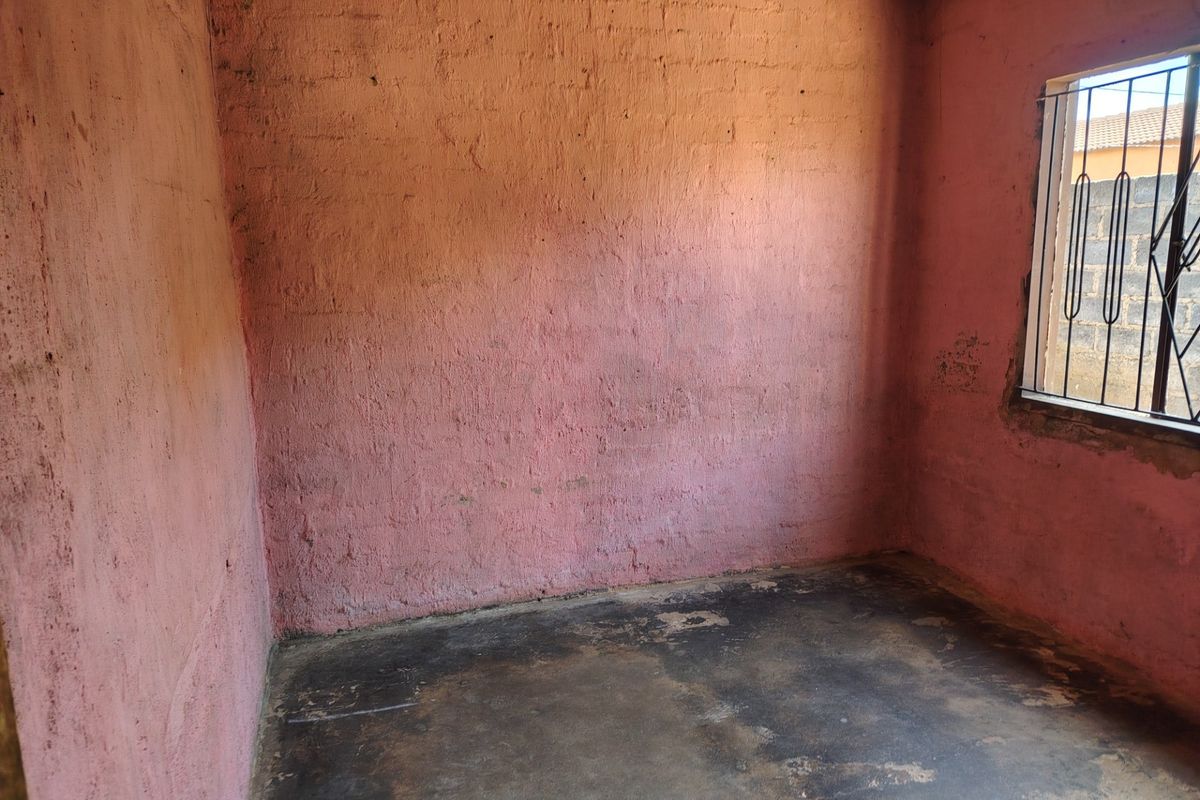 An empty bedroom featuring raw brick walls and a concrete floor, equipped with security bars on the window.