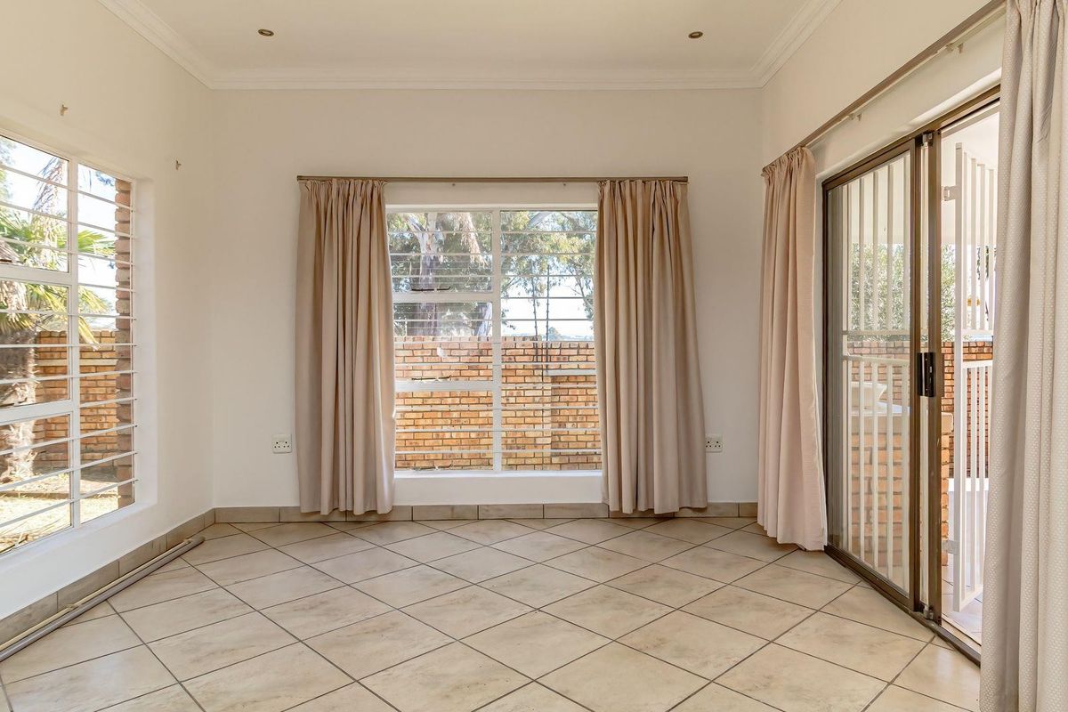 A bright, empty living room featuring neutral tiled flooring, recessed lighting, and large windows with security bars, providing access to the outdoor area.