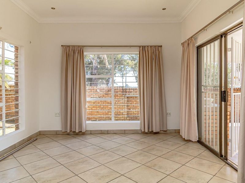 A bright, empty living room featuring neutral tiled flooring, recessed lighting, and large windows with security bars, providing access to the outdoor area.