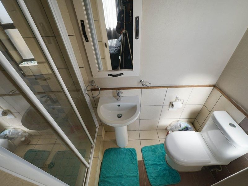 A compact bathroom featuring a pedestal washbasin, a glass-enclosed shower, and a standard toilet with neutral wall and floor tiling.