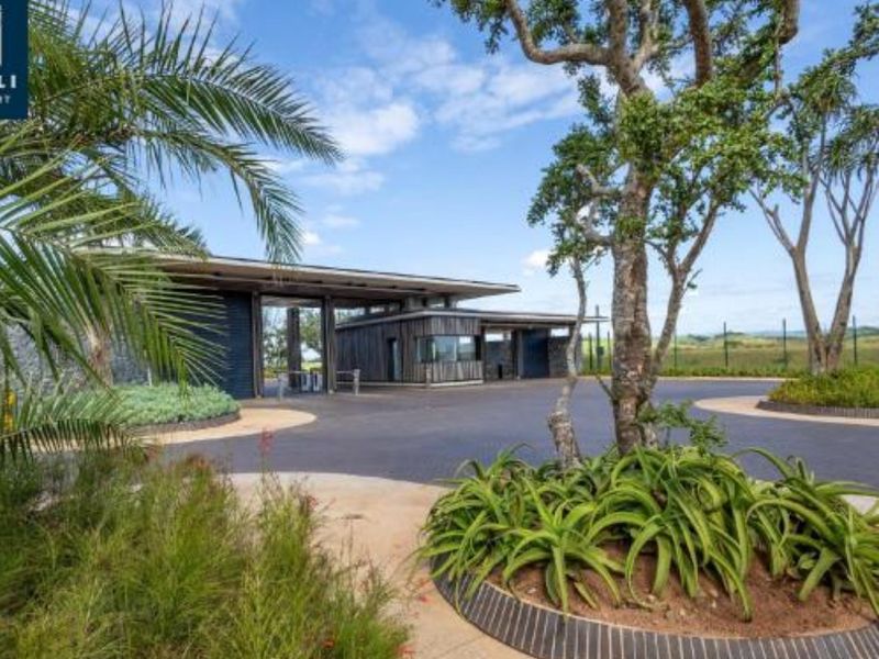 A well-maintained modern security gatehouse entrance featuring a paved driveway and landscaped garden beds with mature trees.