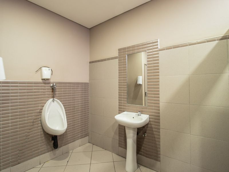 A clean commercial bathroom facility featuring a wall-mounted urinal, a pedestal basin, and neutral ceramic wall and floor tiling.