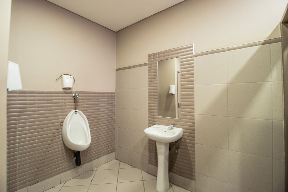 A clean commercial bathroom facility featuring a wall-mounted urinal, a pedestal basin, and neutral ceramic wall and floor tiling.