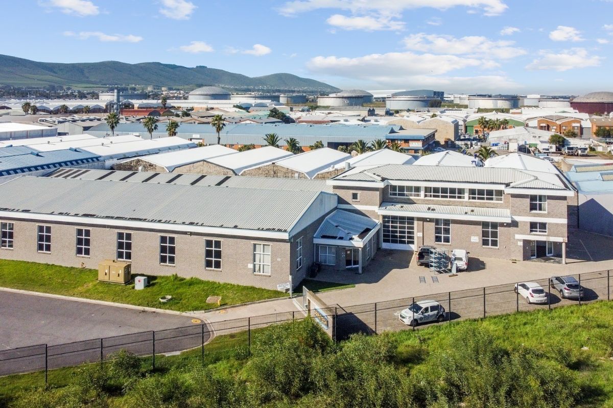 Aerial view of an industrial property featuring a large warehouse complex with metal roofing, brick facade, and secure perimeter fencing in an industrial park.