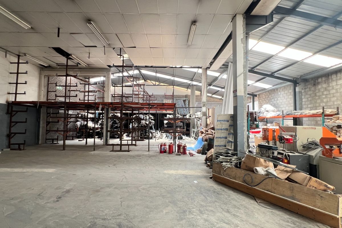 Spacious industrial warehouse area featuring concrete flooring, exposed brick walls, integrated shelving units, and functional overhead fluorescent lighting.