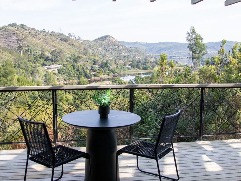 A wooden balcony featuring a decorative metal balustrade, offering expansive views of the surrounding hills, forest, and river landscape.