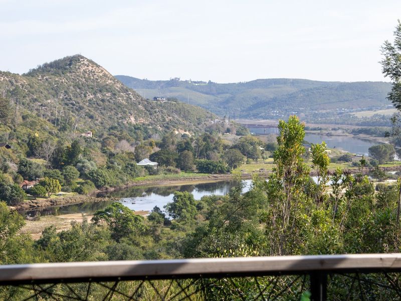 A scenic elevated view overlooking a river, lush valley, and rolling hills, captured from a balcony with a metal railing.