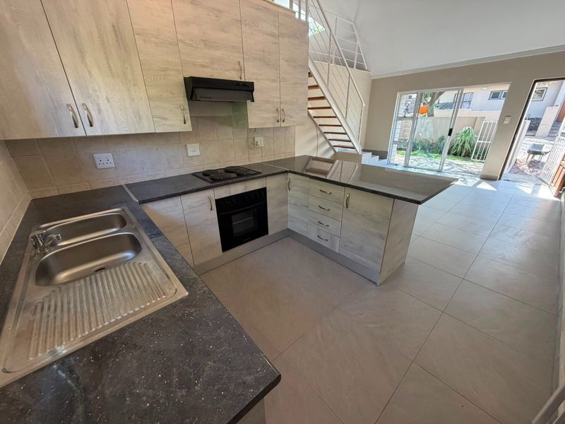 Open-plan kitchen featuring light wood-finish cabinetry, dark countertops, and integrated appliances, flowing into a bright living area with direct access to the rear patio.