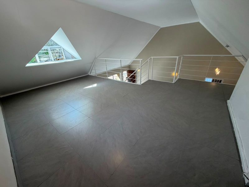 A spacious, empty loft-style living area featuring modern grey floor tiles, a unique triangular window, and a white metal balustrade overlooking the lower level.