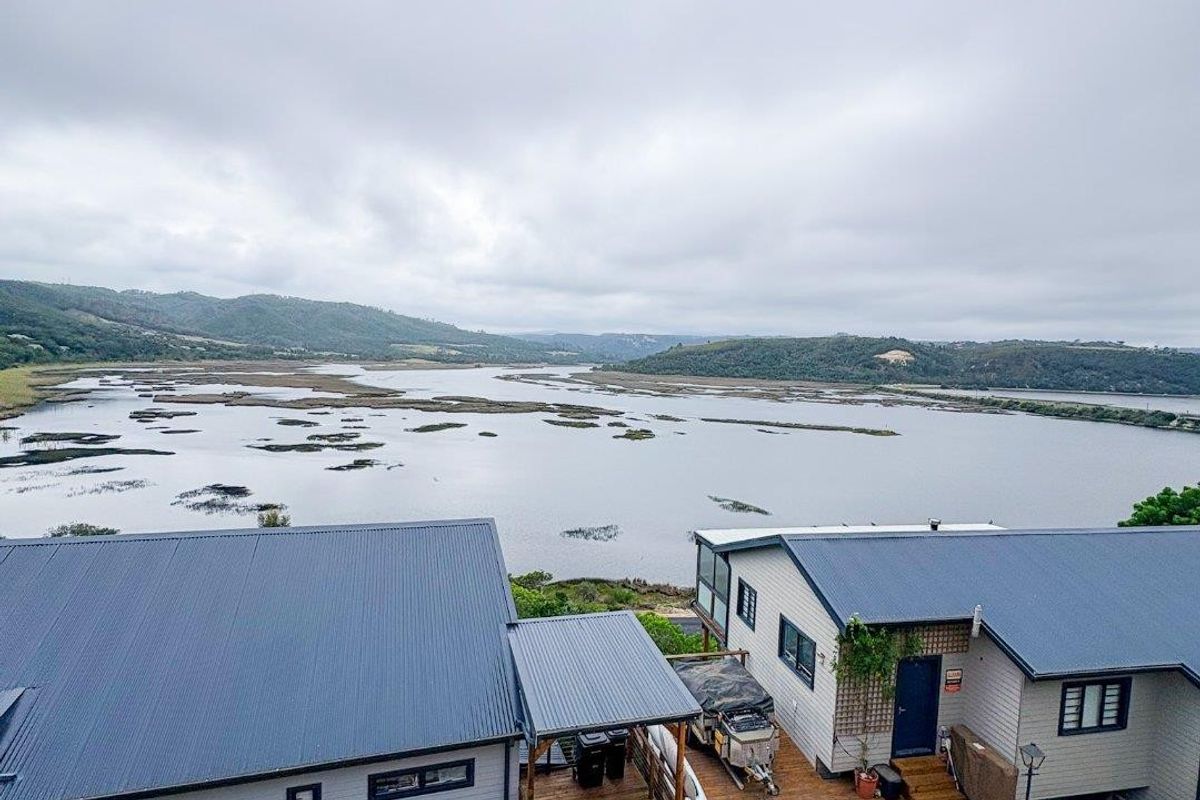 Aerial view of the property featuring metal roofing, timber decking, and a brick paved driveway, overlooking a scenic estuary and surrounding landscape.