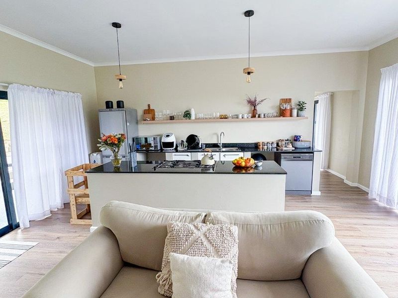 Open-plan living area featuring a modern kitchen with a central island, gas cooktop, floating shelving, and light timber-look flooring.