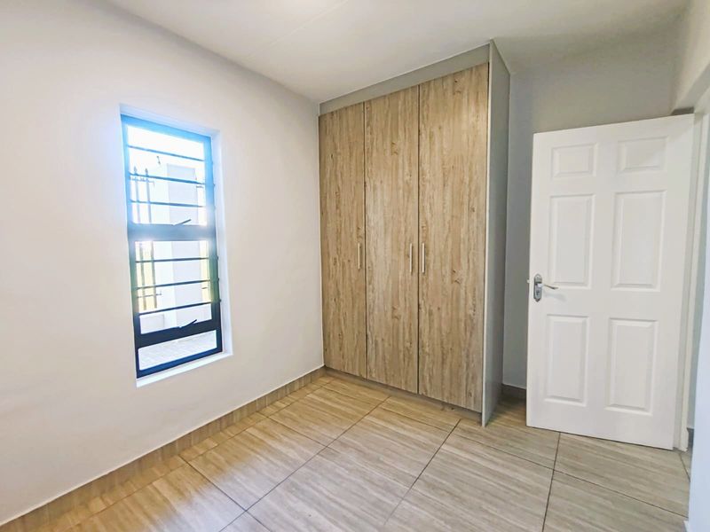 A clean, empty bedroom featuring modern wood-look tiled flooring, a built-in wardrobe with wood-grain finish, and a window fitted with security bars.