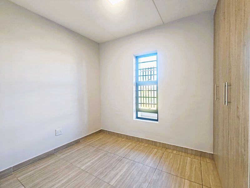 A clean, empty bedroom featuring neutral tiled flooring, built-in wooden cupboards, and a window fitted with security bars.