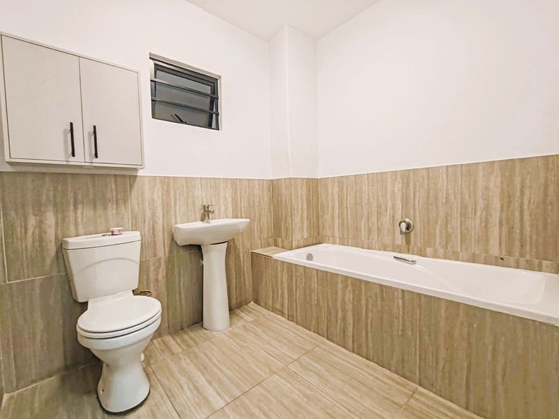 A clean, neutral-toned bathroom featuring a pedestal sink, a built-in bathtub, a toilet, and a wall-mounted storage cabinet with tiled finishes throughout.