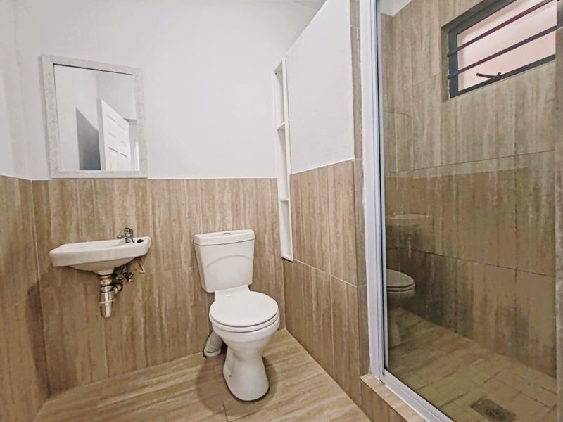 A functional bathroom featuring a wall-mounted basin, toilet, and a tiled shower enclosure with neutral wall and floor tiling.