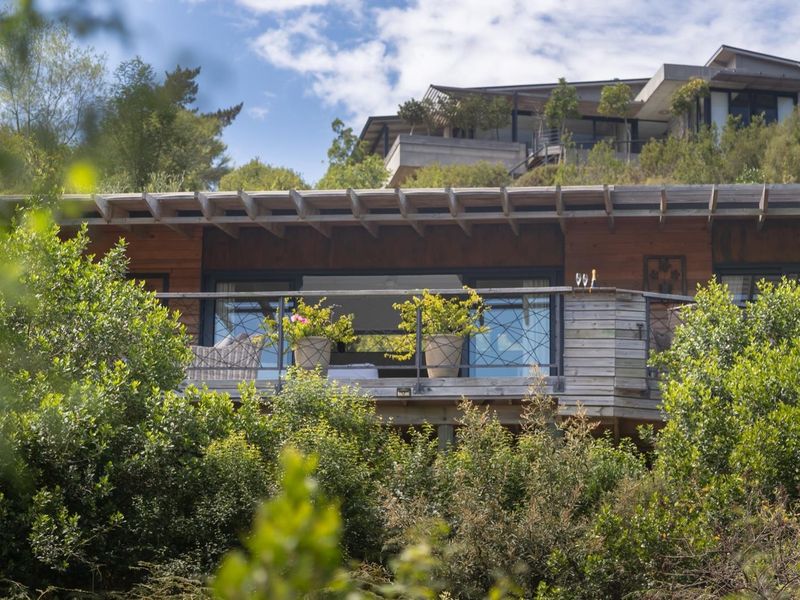 A rear view of the property featuring timber cladding, a wooden pergola, and a balcony with a metal balustrade overlooking lush greenery.