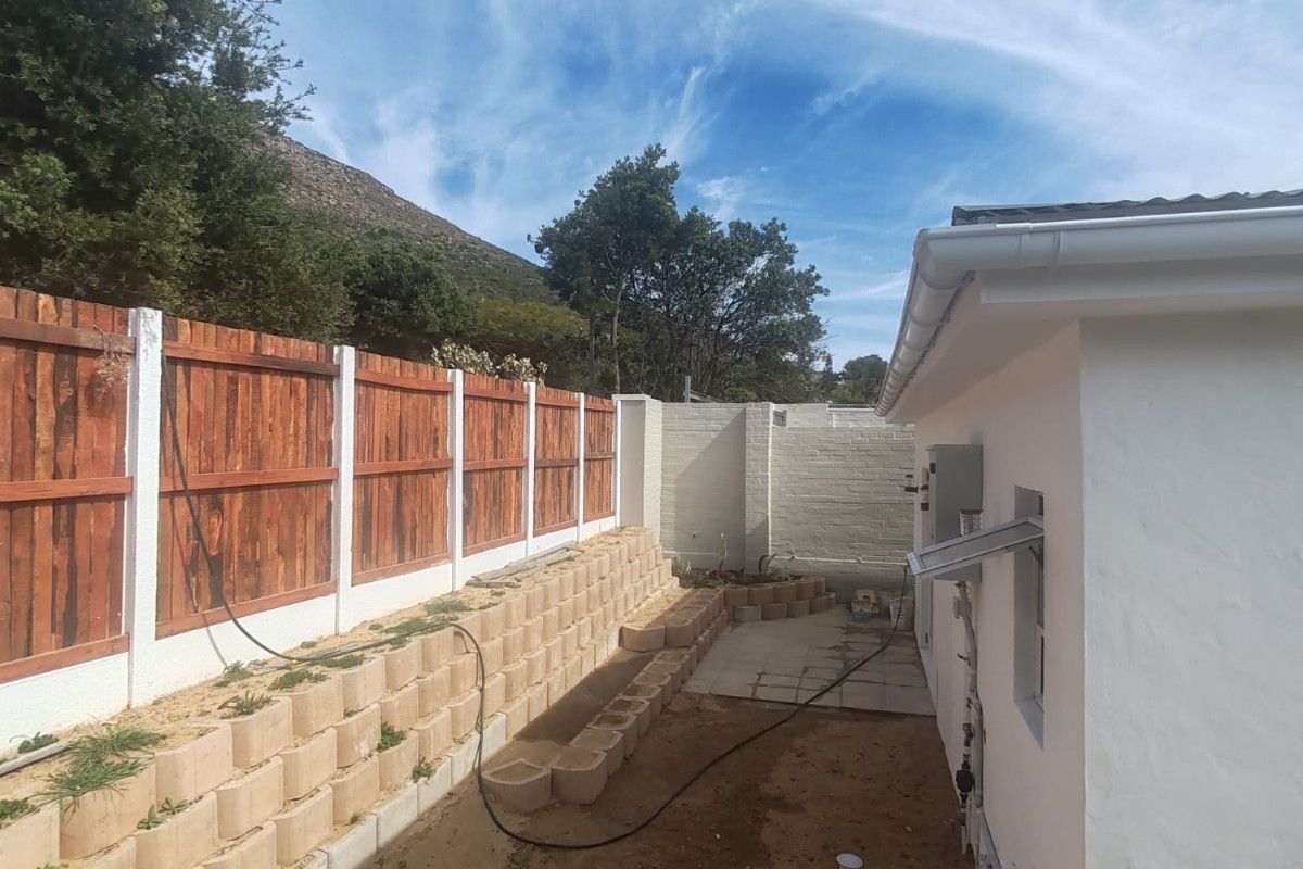 Rear exterior view showing a timber fence, tiered retaining wall, and an unfinished paved patio area adjacent to the house structure.