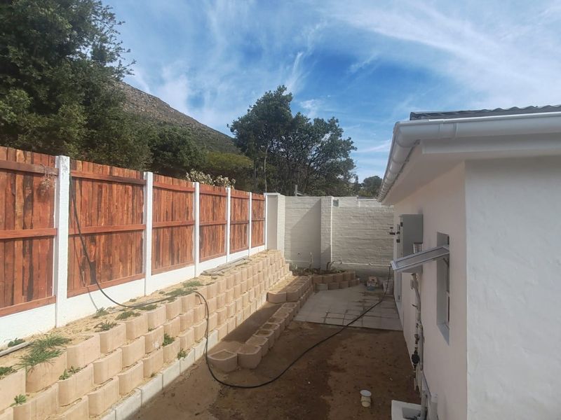 Rear exterior view showing a timber fence, tiered retaining wall, and an unfinished paved patio area adjacent to the house structure.