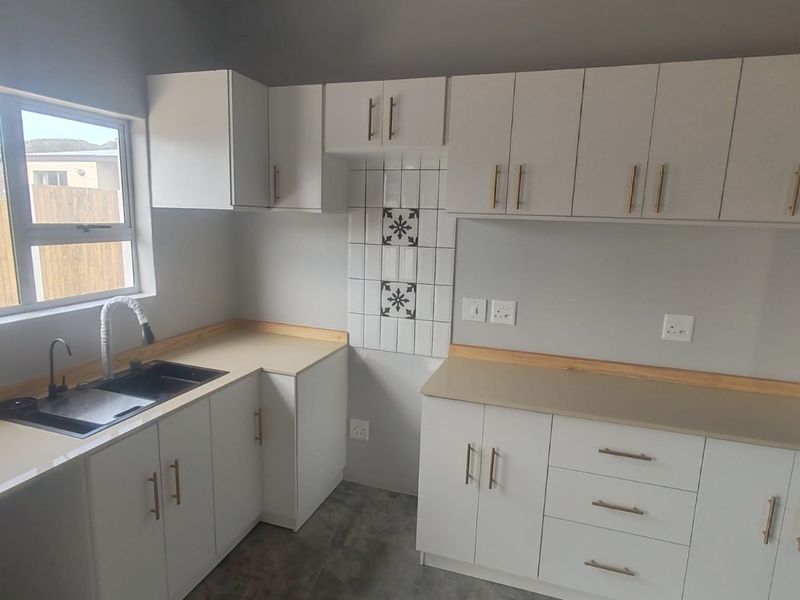 A modern, clean kitchen featuring white cabinetry, light-toned countertops, a double sink, and a decorative tiled splashback under an exposed timber beam.