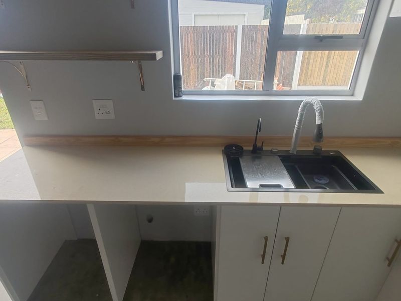 A modern kitchen area featuring a clean quartz countertop, undermount sink with gooseneck faucet, white cabinetry, and floating timber shelving.