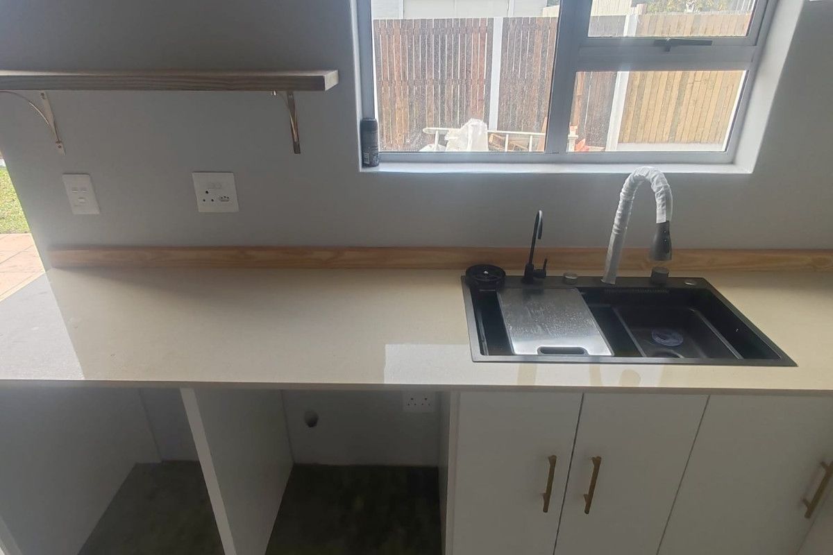 A modern kitchen area featuring a clean quartz countertop, undermount sink with gooseneck faucet, white cabinetry, and floating timber shelving.