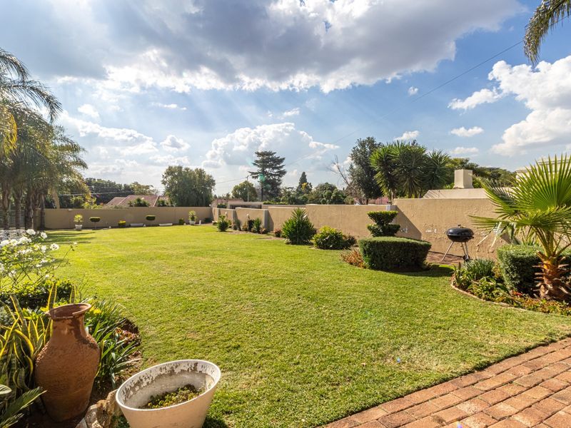 A spacious, well-maintained backyard featuring a large lawn, perimeter boundary walls, brick paving, and a built-in outdoor fireplace.