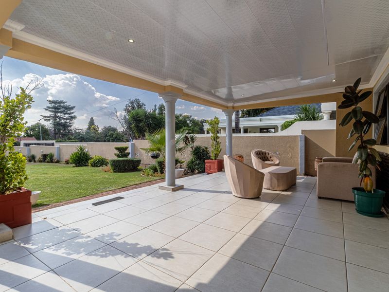 A spacious covered patio featuring tiled flooring, recessed ceiling lights, and decorative support columns, overlooking a well-maintained garden area.