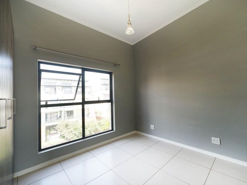 A clean, empty bedroom featuring neutral grey walls, large aluminium-framed windows, tiled flooring, and a built-in cupboard.