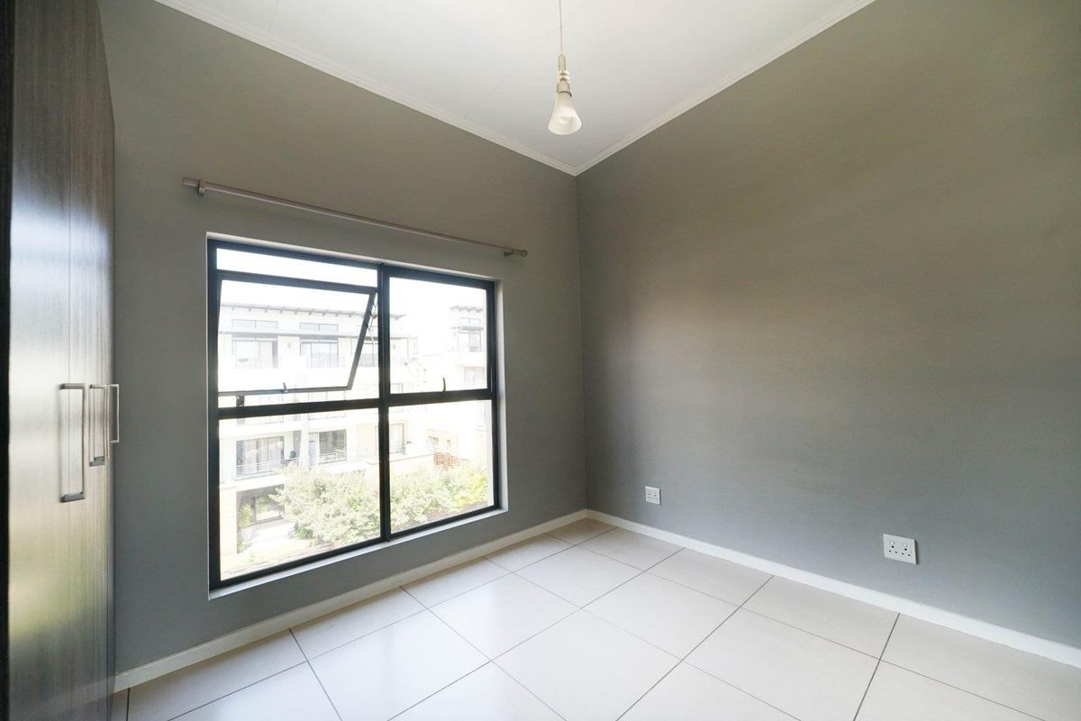 A clean, empty bedroom featuring neutral grey walls, large aluminium-framed windows, tiled flooring, and a built-in cupboard.