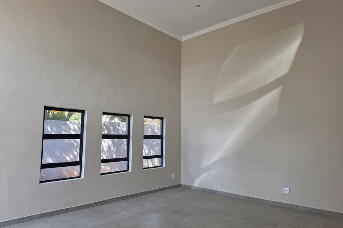 A bright, empty living area featuring neutral walls, tiled flooring, and three modern aluminium-framed windows that allow for natural light.