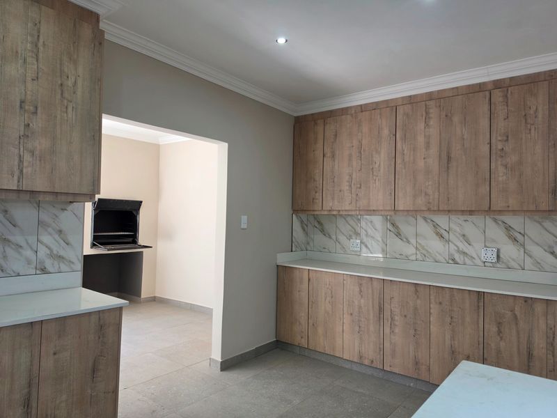 Modern kitchen featuring wood-look cabinetry, stone countertops, and marble-look backsplash, with an adjacent room containing a built-in braai.