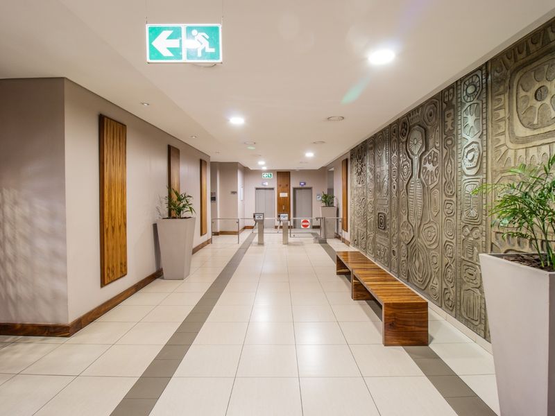 A modern commercial building lobby featuring tiled flooring, a decorative textured feature wall, elevator access, and secure turnstile entry points.
