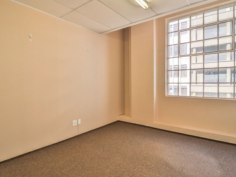 A vacant office space featuring carpeted flooring, a suspended ceiling with integrated lighting, and a large glass block window providing natural light.
